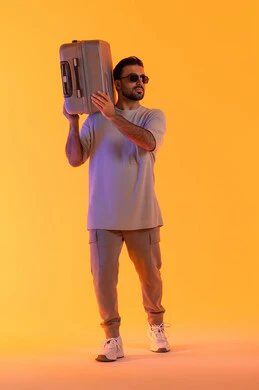 Tourism during the summer vacation, gestures of joy and happiness, preparing to spend an enjoyable summer trip, going on recreational tours, a portrait of an Emirati Gulf Arab man wearing casual attire and sunglasses, carrying a luggage bag on his shoulder, full-body shot, orange background.
