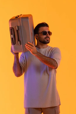 نظرات الثقة والرجولة، مفهوم السياحة في إجازة الصيف، ايماءات السرور والفرح، التحضير قضاء رحلة صيفية ممتعة، القيام بالجولات الترفيهية، بورتريه مقرب لرجل عربي خليجي اماراتي يرتدي الزي الكاجوال ونظارة شمسية يحمل على كتفه حقيبة الامتعة، خلفية برتقاليه