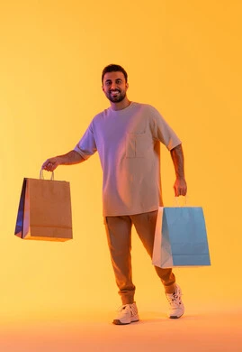 التسوق وشراء الحاجيات والهدايا، الاستفادة من العروضات والتخفيضات، النظر إلى الكاميرا بإيماءات البهجة والسرور، بورتريه لرجل عربي خليجي إماراتي مبتسم يرتدي الزي الكاجوال يحمل بيديه أكياس ورقية، الترويج للمنتجات والماركات العالمية، خلفية برتقاليه