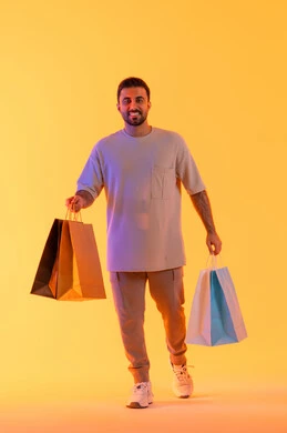 شراء الحاجيات والهدايا، الاستفادة من العروضات والتخفيضات، النظر إلى الكاميرا بإيماءات البهجة والسرور، بورتريه لرجل عربي خليجي إماراتي مبتسم يرتدي الزي الكاجوال يحمل بيديه أكياس ورقية، الترويج للمنتجات والماركات العالمية، خلفية برتقاليه