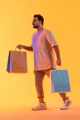 مفهوم التسوق الالكتروني, الاستفادة من العروضات والتخفيضات، التسوق وشراء الحاجيات والهدايا، إيماءات البهجة والسرور، بورتريه لرجل عربي خليجي إماراتي مبتسم يرتدي الزي الكاجوال يحمل بيديه أكياس ورقية، الترويج للمنتجات والماركات العالمية، خلفية برتقاليه