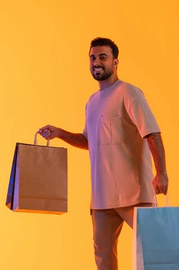 شراء المنتجات و الدفع عن بعد, الاستفادة من العروضات والتخفيضات، التسوق وشراء الحاجيات والهدايا، إيماءات البهجة والسرور، بورتريه لرجل عربي خليجي إماراتي مبتسم يرتدي الزي الكاجوال يحمل بيديه أكياس ورقية، الترويج للمنتجات والماركات العالمية، خلفية برتقاليه