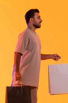 تعابير البهجة والسرور، شراء المنتجات و الدفع عن بعد, الاستفادة من العروضات والتخفيضات، التسوق وشراء الحاجيات والهدايا، بورتريه جانبي لرجل عربي خليجي إماراتي مبتسم يرتدي الزي الكاجوال يحمل بيديه أكياس ورقية، الترويج للمنتجات والماركات العالمية، خلفية برتقاليه