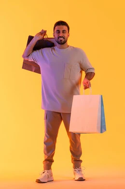 Promoting global products and brands, expressions of joy and happiness, purchasing products and paying remotely, benefiting from offers and discounts, shopping and buying necessities and gifts, a portrait of a smiling Emirati Gulf Arab man wearing casual attire holding paper bags in his hands, with an orange background.