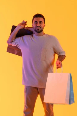 الماركات العالمية والترويج للمنتجات، تعابير البهجة والسرور، شراء المنتجات و الدفع عن بعد, الاستفادة من العروضات والتخفيضات، التسوق وشراء الحاجيات والهدايا، بورتريه لرجل عربي خليجي إماراتي مبتسم يرتدي الزي الكاجوال يحمل بيديه أكياس ورقية، خلفية برتقاليه