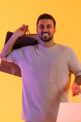 التنزيلات والحسومات الموسمية، تعابير البهجة والسرور، شراء المنتجات و الدفع عن بعد, الاستفادة من العروضات والتخفيضات، التسوق وشراء الحاجيات والهدايا، بورتريه لرجل عربي خليجي إماراتي مبتسم يرتدي الزي الكاجوال يحمل بيديه أكياس ورقية، خلفية برتقاليه