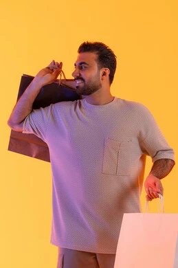 الحسومات والتنزيلات الموسمية، تعابير البهجة والسرور، شراء المنتجات و الدفع عن بعد, الاستفادة من العروضات والتخفيضات، التسوق وشراء الحاجيات والهدايا، بورتريه لرجل عربي خليجي إماراتي مبتسم يرتدي الزي الكاجوال يحمل بيديه أكياس ورقية، خلفية برتقاليه