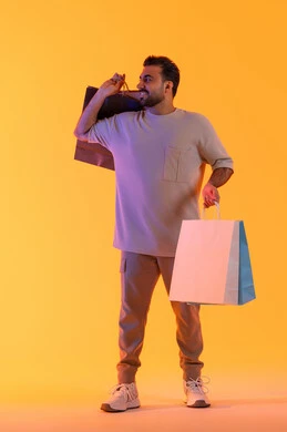 العروض والتخفيضات على كافة الأصناف، تعابير البهجة والسرور، شراء المنتجات و الدفع عن بعد, الاستفادة من العروضات والتخفيضات، التسوق وشراء الحاجيات والهدايا، بورتريه لرجل عربي خليجي إماراتي مبتسم يرتدي الزي الكاجوال يحمل بيديه أكياس ورقية، خلفية برتقاليه