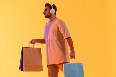 اقتناء المستلزمات والهدايا, تعابير البهجة والسرور، شراء المنتجات و الدفع عن بعد, الاستفادة من العروضات والتخفيضات، التسوق وشراء الحاجيات والهدايا، بورتريه لرجل عربي خليجي إماراتي مبتسم يرتدي الزي الكاجوال وسماعة الرأس يحمل بيديه أكياس ورقية، خلفية برتقاليه