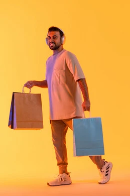 الاستماع غلى المقاطع الصوتية، تعابير البهجة والسرور، شراء المنتجات و الدفع عن بعد, الاستفادة من العروضات والتخفيضات، التسوق وشراء الحاجيات والهدايا، بورتريه لرجل عربي خليجي إماراتي مبتسم يرتدي الزي الكاجوال وسماعة الرأس يحمل بيديه أكياس ورقية، خلفية برتقاليه