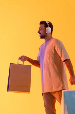 Shopping experience, expressions of joy and happiness, purchasing products and paying remotely, benefiting from offers and discounts, shopping for necessities and gifts, a portrait of a smiling Emirati Gulf Arab man wearing casual attire and headphones, holding paper bags in his hands, with an orange background.