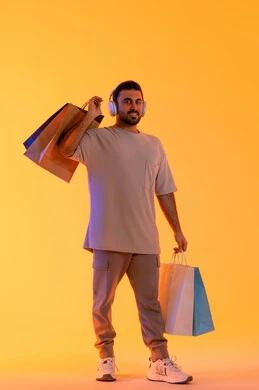 الترفيه والتسيلة خلال التسوق، تعابير البهجة والسرور، شراء المنتجات و الدفع عن بعد, الاستفادة من العروضات والتخفيضات، التسوق وشراء الحاجيات والهدايا، بورتريه لرجل عربي خليجي إماراتي مبتسم يرتدي الزي الكاجوال وسماعة الرأس يحمل بيديه أكياس ورقية، خلفية برتقاليه