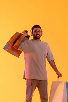 التواصل والتسيلة خلال التسوق، تعابير البهجة والسرور، شراء المنتجات و الدفع عن بعد, الاستفادة من العروضات والتخفيضات، التسوق وشراء الحاجيات والهدايا، بورتريه لرجل عربي خليجي إماراتي مبتسم يرتدي الزي الكاجوال وسماعة الرأس يحمل بيديه أكياس ورقية، خلفية برتقاليه