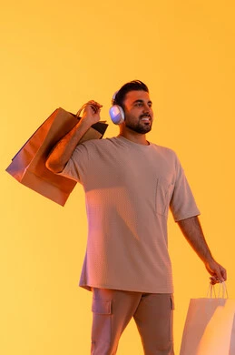Enjoying shopping at the mall, expressions of joy and happiness, purchasing products and paying remotely, benefiting from offers and discounts, shopping for necessities and gifts, a portrait of a smiling Emirati Gulf Arab man wearing casual attire and headphones, holding paper bags in his hands, with an orange background.