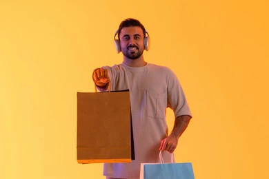 عرض كيس ورقي بني، تعابير البهجة والسرور، شراء المنتجات و الدفع عن بعد, الاستفادة من العروضات والتخفيضات، التسوق وشراء الحاجيات والهدايا، بورتريه لرجل عربي خليجي إماراتي مبتسم يرتدي الزي الكاجوال وسماعة الرأس يحمل بيديه أكياس ورقية، خلفية برتقاليه