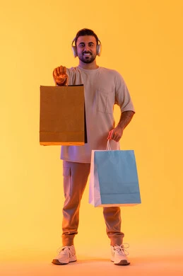 التبضع وأخذ الحاجات اللازمة، تعابير البهجة والسرور، شراء المنتجات و الدفع عن بعد, الاستفادة من العروضات والتخفيضات، التسوق وشراء الحاجيات والهدايا، بورتريه لرجل عربي خليجي إماراتي مبتسم يرتدي الزي الكاجوال وسماعة الرأس يحمل بيديه أكياس ورقية، خلفية برتقاليه