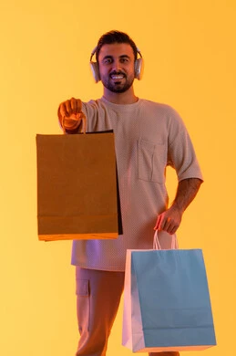 Commercial purchases and gifts, expressions of joy and happiness, buying products and paying remotely, benefiting from offers and discounts, shopping and buying necessities and gifts, a portrait of a smiling Emirati Gulf Arab man wearing casual attire and headphones holding paper bags in his hands, with an orange background.
