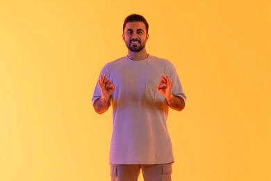 Achieving goals and ambitions, the concept of attractiveness and self-confidence, standing upright and straight, looking with gestures of joy and comfort, a portrait of a smiling Emirati Gulf Arab man wearing casual attire raising his hands in a perfect or complete shape, the concept of masculinity and elegance, with an orange background.