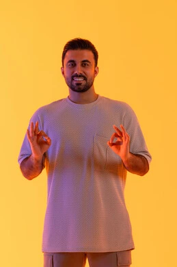 الإشارة إلى تحقيق هدف محدد، مفهوم الوسامة والثقة بالنفس، الوقوف باستقامة واعتدال, النظر بإيماءات السرور والراحة، بورتريه لرجل عربي خليجي اماراتي مبتسم يرتدي الزي الكاجوال يرفع يديه على شكل بيرفكت أو تمام، مفهوم الرجولة والاناقة، خلفية برتقاليه