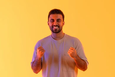 The feeling of strength and success, expressions of interaction and engagement, a close-up portrait of a young Arab Gulf Emirati man wearing casual attire raising his hands with palms together, a Saudi man looking with gestures of happiness, victory, and triumph, clubs and cheering for the favorite team, yellow background.