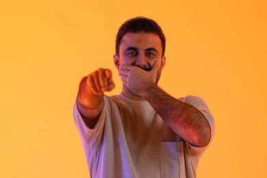 مفهوم كتم الاسرار، تعابير السرور والسعادة، الاشارة بإصبع السبابة إلى شيء ما، بورتريه لشاب عربي خليجي  اماراتي يرتدي الزي الكاجوال يضع يده على فمه، رجل سعودي يقوم بالنظر إلى احد ما، خلفية صفراء