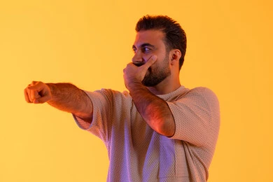 مفهوم كتم الاسرار، تعابير السرور والسعادة، الاشارة بإصبع السبابة إلى شيء ما، بورتريه لشاب عربي خليجي  اماراتي يرتدي الزي الكاجوال يضع يده على فمه، رجل سعودي يقوم بالنظر إلى احد ما، خلفية صفراء