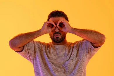 The concept of future planning for goals and ambitions, a love for adventure and exploration, a close-up portrait of a young Arab Gulf Emirati man wearing casual attire, placing his hand over his eye in the shape of a telescope, a Saudi man looking at distant things, with a yellow background.