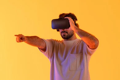 رؤية المشاهد الخيالية، الاشارة بإصبع السبابة إلى شيء ما، استخدام الأجهزة التقنية الحديثة، التفاعل والاندماج مع الواقع الافتراضي، بورتريه مقرب لشاب عربي خليجي إماراتي يرتدي الزي الكاجوال يضع نظارات الفي آر، رجل سعودي يقضي أوقات الفراغ بشكل ممتع، خلفية صفراء