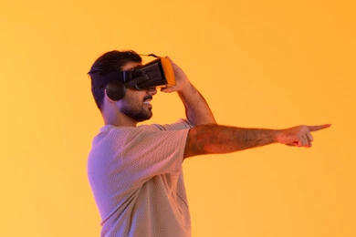 Seeing imaginary scenes, pointing with the index finger at something, using modern technological devices, interacting and merging with virtual reality, a close-up portrait of a young Emirati Gulf Arab man wearing casual attire and VR glasses, a Saudi man enjoying his leisure time, yellow background.
