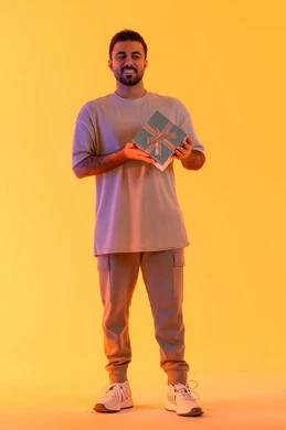 التعبير عن الحب والتقدير والاحترام، تقديم الهدايا بالاعياد والمناسبات الخاصة، مفهوم الشكر والامتنان، رجل سعودي ينظر إلى الكاميرا بايماءات الفرح والسرور، بورتريه لشاب عربي خليجي اماراتي يرتدي الزي الكاجوال يحمل بيديه صندوق هدية، الجسم بالطول الكامل، خلفية صفراء
