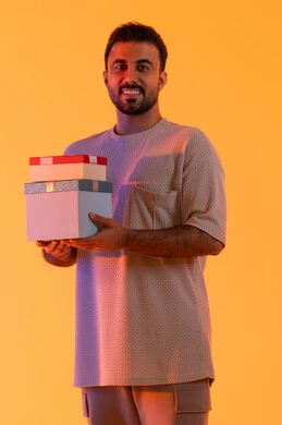 مفهوم الشكر والامتنان، التعبير عن الحب والتقدير والاحترام، تقديم الهدايا بالاعياد والمناسبات الخاصة، رجل سعودي ينظر إلى الكاميرا بايماءات الفرح والسرور، بورتريه لشاب عربي خليجي اماراتي يرتدي الزي الكاجوال يحمل بيديه صندوق هدية، خلفية صفراء