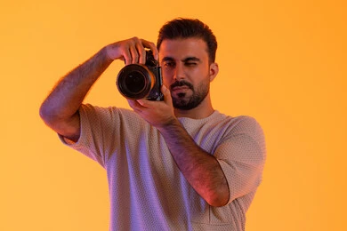 استخدام كاميرات رقمية عالية الدقة, ممارسة هواية التصوير بتعابير السرور, اتقان التصوير الفوتوغرافي، مصور سعودي محترف يقوم بالتقاط صور تذكارية, بورتريه مقرب لشاب عربي خليجي اماراتي يرتدي الزي الكاجوال يمسك بيده كاميرا فوتوغرافية، خلفية صفراء