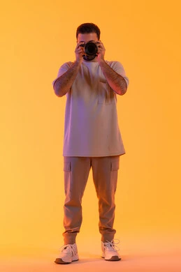 اتقان التصوير الفوتوغرافي باستخدام كاميرات رقمية عالية الدقة, مصور سعودي محترف يقوم بالتقاط صور تذكارية, ممارسة هواية التصوير بتعابير السرور, بورتريه لشاب عربي خليجي اماراتي يرتدي الزي الكاجوال يمسك بيده كاميرا فوتوغرافية، صورة الجسم بالكامل، خلفية صفراء