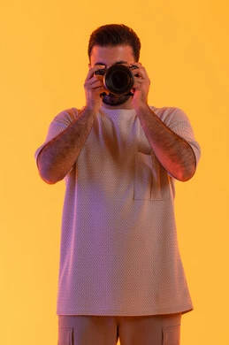 Practicing the hobby of photography with expressions of joy, mastering photography using high-resolution digital cameras, a professional Saudi photographer capturing commemorative photos, a portrait of a young Arab Gulf Emirati man wearing casual attire holding a camera in his hand, with a yellow background.