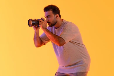 Practicing the hobby of photography with expressions of joy, mastering photography using high-resolution digital cameras, a professional Saudi photographer capturing commemorative photos, a portrait of a young Arab Gulf Emirati man wearing casual attire holding a camera in his hand, with a yellow background.