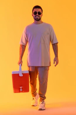 الاستمتاع بالمشروبات الباردة وقضاء وقت ممتع في العطلة الصيفية, السياحة في إحدى منتجعات السعودية, بورتريه لشاب عربي خليجي سعودي مبتسم يرتدي الزي الكاجوال والنظارة الشمسية، رجل اماراتي يحمل حافظة العصائر وينظر إلى الكاميرا, صورة الجسم بالكامل, خلفية صفراء