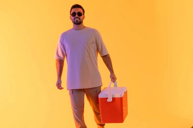 السياحة في إحدى منتجعات الإمارات, الاستمتاع بالمشروبات الباردة وقضاء وقت ممتع في العطلة الصيفية, بورتريه لشاب عربي خليجي سعودي مبتسم يرتدي الزي الكاجوال والنظارة الشمسية، رجل اماراتي يحمل حافظة العصائر وينظر إلى الكاميرا, خلفية صفراء