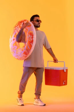 قضاء وقت ممتع في فصل الصيف, بورتريه لشاب عربي خليجي اماراتي مبتسم يرتدي الزي الكاجوال والنظارة الشمسي, رجل سعودي يحمل العوامة يستعد للسباحة في الشاطئ, حافظة البرودة للعصائر, الاستمتاع بالعطلة الصيفية, الجسم بالطول الكامل, خلفية صفراء