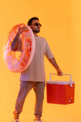 قضاء وقت ممتع في فصل الصيف, بورتريه لشاب عربي خليجي اماراتي مبتسم يرتدي الزي الكاجوال والنظارة الشمسي, رجل سعودي يحمل العوامة يستعد للسباحة في الشاطئ, حافظة البرودة للعصائر, الاستمتاع بالعطلة الصيفية, الجسم بالطول الكامل, خلفية صفراء