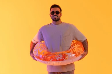 النظر إلى الكاميرا بتعابير السرور، قضاء أوقات صيفية سياحية, الاستمتاع في شواطئ الامارات, التسلية في إحدى المنتجعات الترفيهيه, بورتريه لشاب عربي خليجي سعودي مبتسم يرتدي الزي الكاجوال والنظارة، رجل اماراتي يمسك عوامة السباحة,  خلفية صفرء