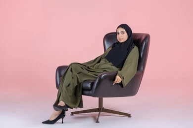 The concept of femininity and self-confidence, a portrait of an Emirati Gulf Arab girl wearing a hijab and an abaya sitting on a chair looking at the camera, attention to elegance and appearance, gestures of joy and happiness, pink background.