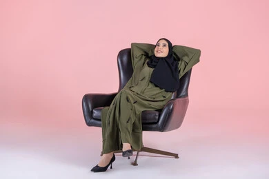 The feeling of tiredness and lethargy and the need for sleep, gestures of comfort and relaxation, looking up at something, a portrait of an Arab Gulf Emirati woman wearing a hijab and an abaya sitting on a chair with her hands behind her head, expressing contemplation and joy, with a pink background.