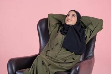 إيماءات الراحة والإسترخاء، النظر للأعلى إلى شيء ما، الشعور بالتعب و الخمول والحاجة إلى النوم, بورتريه لامرأة عربيه خليجيه اماراتية محجبه ترتدي العباءة تجلس على كرسي تضع يديها وراء رأسها بتعابير التأمل والسرور، خلفية وردية