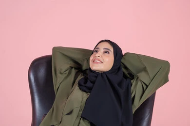 Looking up at something, gestures of comfort and relaxation, feeling tired and lethargic and the need for sleep, a close-up portrait of an Arab Gulf Emirati woman wearing a hijab and an abaya sitting on a chair with her hands behind her head, expressing contemplation and joy, with a pink background.