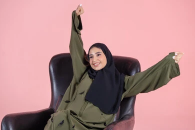 A hand movement indicating relaxation, expressions of happiness and joy, a portrait of a Gulf Arab Emirati woman wearing a hijab and an abaya sitting on a chair and stretching her body, various facial and hand gestures, pink background.