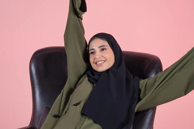 تعابير السعادة و السرور، بورتريه لإمرأة عربية خليجية اماراتية محجبه ترتدي العباءة تجلس على الكرسي وتقوم بتمديد جسدها، ايماءات وجه ويدين مختلفة، حركة يدين تدل على الاسترخاء، خلفية وردية