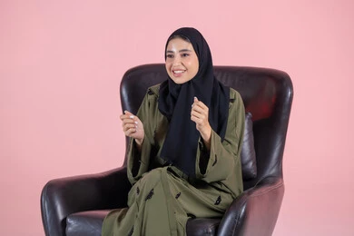 Facial and hand gestures indicating encouragement and enthusiasm, a portrait of a Gulf Arab Emirati woman wearing a hijab and an abaya sitting on a chair celebrating victory and triumph, happiness in winning and achieving goals, pink background.