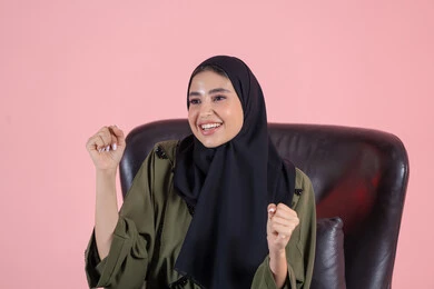 السعادة بالفوز و تحقيق الاهداف، ايماءات وجه ويدين تدل على التشجيع والحماس، بورتريه مقرب لامرأة عربية خليجية اماراتية محجبة ترتدي العباءة تجلس على كرسي وتحتفل بالفوز والانتصار، خلفية وردية