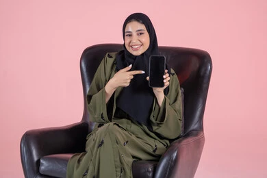 Pointing with the index finger at the mobile phone, displaying mobile applications, looking at the camera with gestures of happiness and joy, showing a blank black screen, a portrait of an Emirati Gulf Arab woman wearing a hijab and an abaya sitting on a chair holding a mobile phone in her hand, using a modern and advanced technological device, pink background.