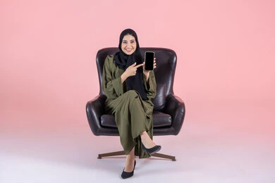 Pointing the index finger towards the mobile phone, displaying mobile applications, looking at the camera with gestures of happiness and joy, showing a blank black screen, a portrait of a Gulf Arab Emirati woman wearing a hijab and an abaya sitting on a chair holding a mobile phone in her hand, using a modern and advanced technological device, pink background.